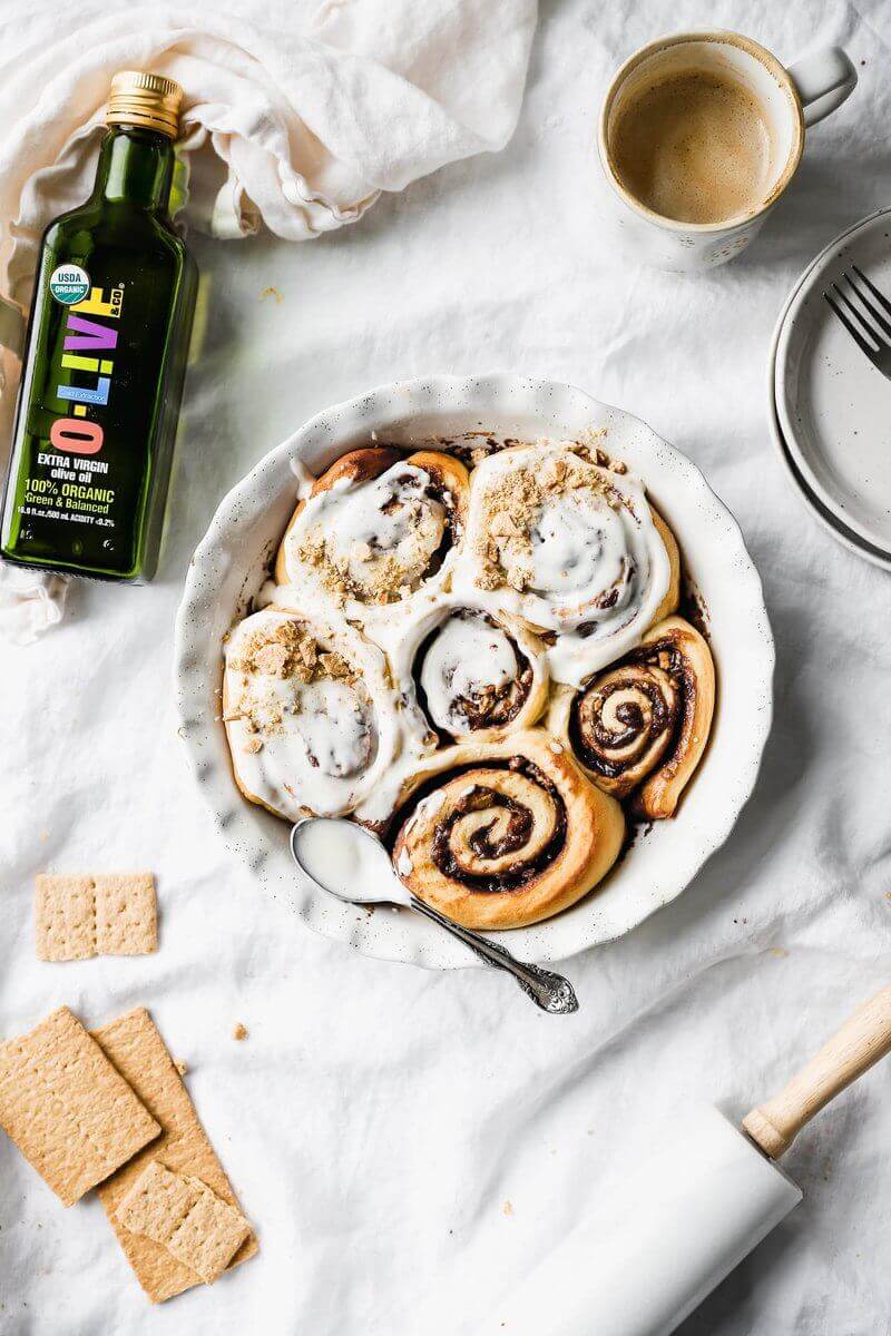 Recipiente blanco con Cinnamon rolls junto a una botella abierta de 500ml de aceite de oliva O-Live & Co.
