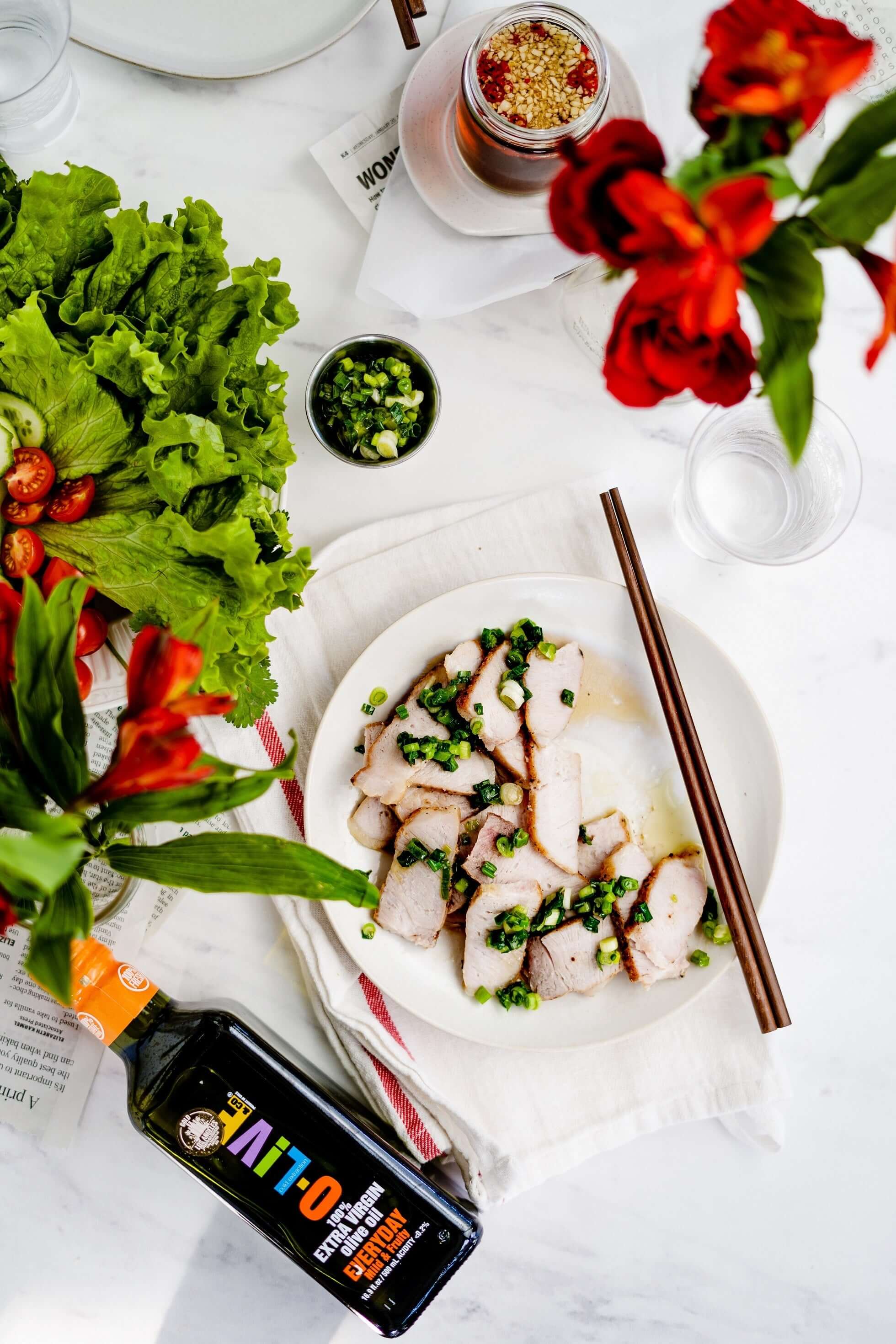 Un plato con chuletas de cerdo vietnamitas junto a dos floreros, una fuente con verduras y una botella de aceite de oliva O-Live & Co.