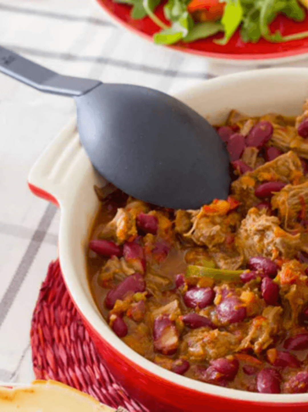 Recipiente rojo que tiene chili y carne condimentado con aceite de oliva O-Live & Co, con una cuchara gris en su interior.