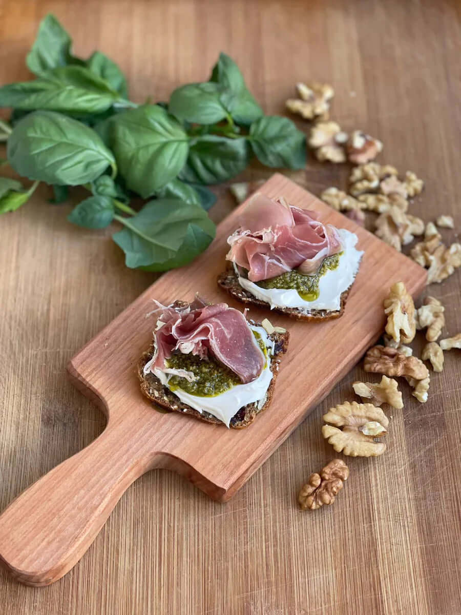 Dos tostaditas con queso crema, pesto y jamón serrano sobre una tabla de madera junto a un montón de nueces y hojas de albahaca.