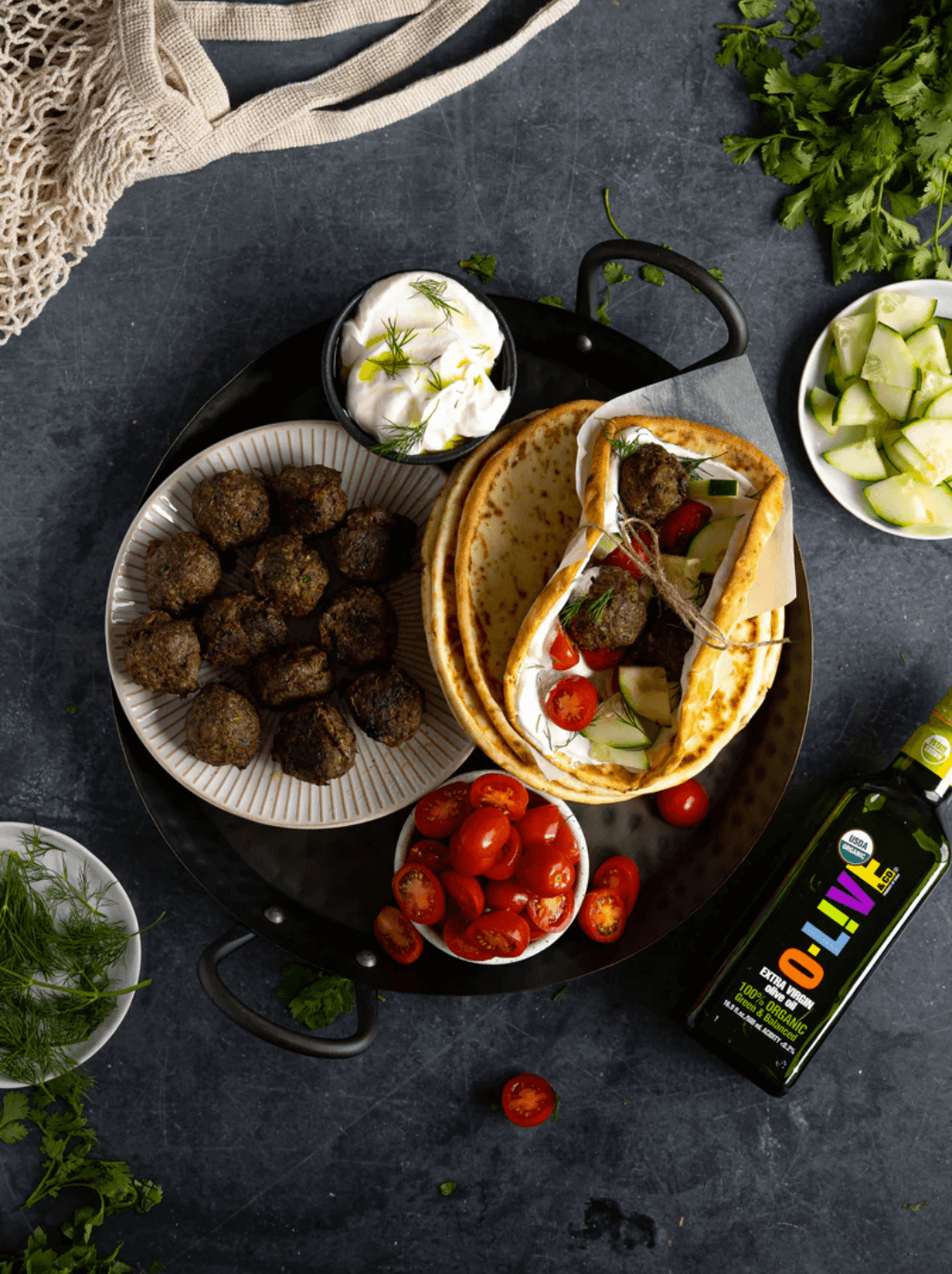 Bandeja negra con albóndigas mediterráneas, queso, tomates cherrys, pepinos cortados y pitas. Todo junto a una botella de 500ml de aceite de oliva O-Live & Co.